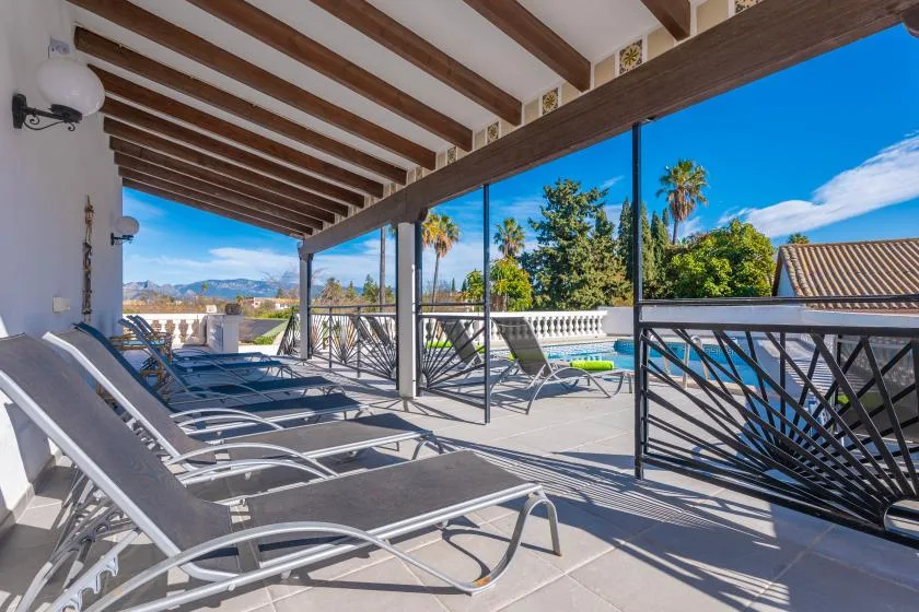 A covered outdoor terrace with lounge seating overlooks a pool and distant hills.
