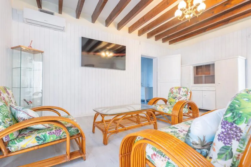 A bright living room with rattan seating, a glass coffee table, and a wall-mounted television.