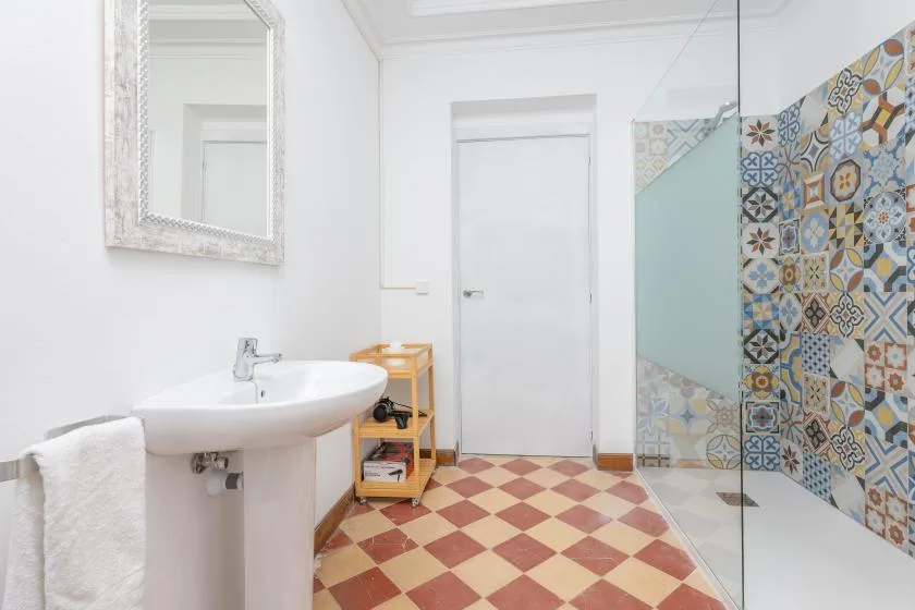 A bright bathroom has a pedestal sink, patterned tile, and a glass shower screen.