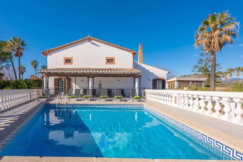 A swimming pool sits in front of a white house with a shaded patio.