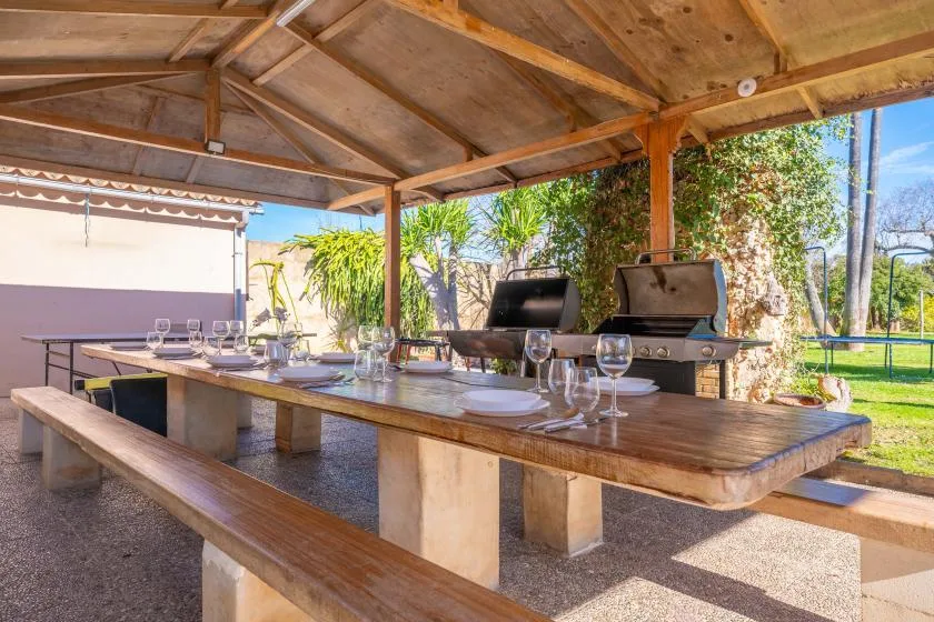A covered outdoor dining area with a long table and a barbecue grill.