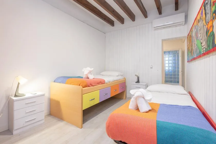 A bright bedroom with colorful beds, a small dresser, and exposed ceiling beams.