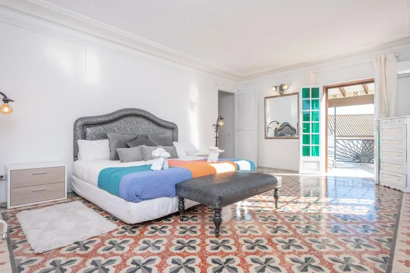 A bright bedroom with a bed, a padded bench, and patterned tile flooring.
