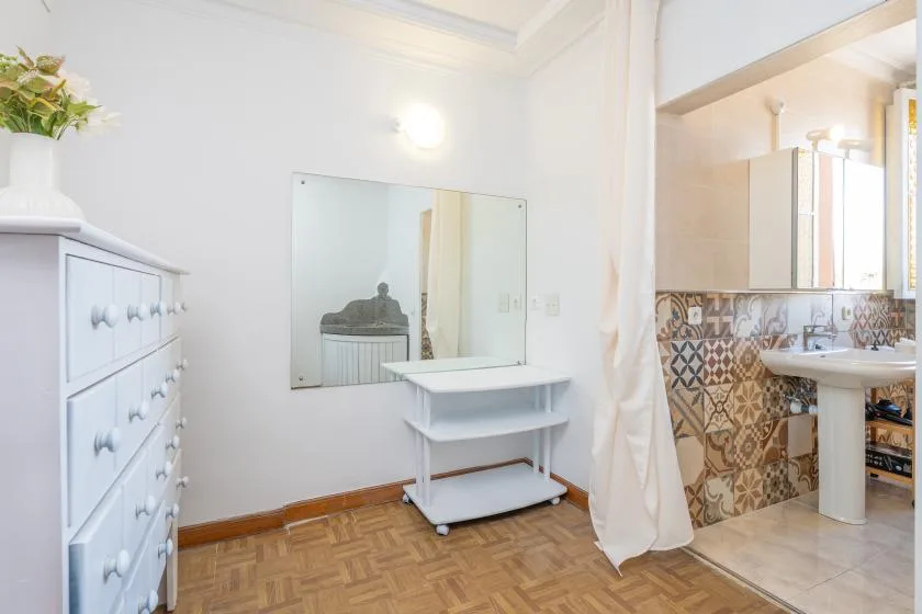 A bright bedroom area has a dresser, mirror, and small table beside an adjoining bathroom.