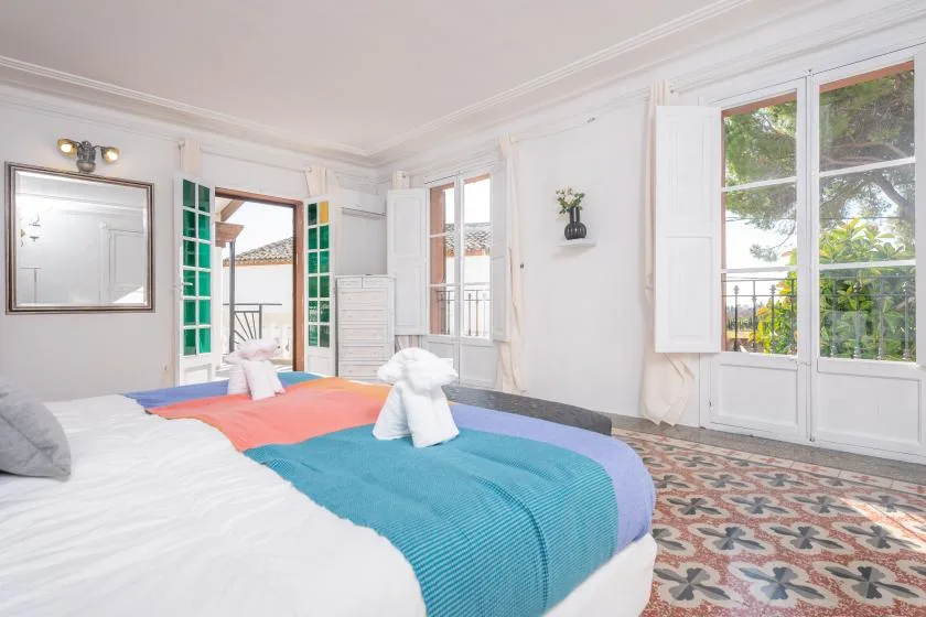 A bright bedroom with colorful bedding and tall windows opening onto a leafy view.