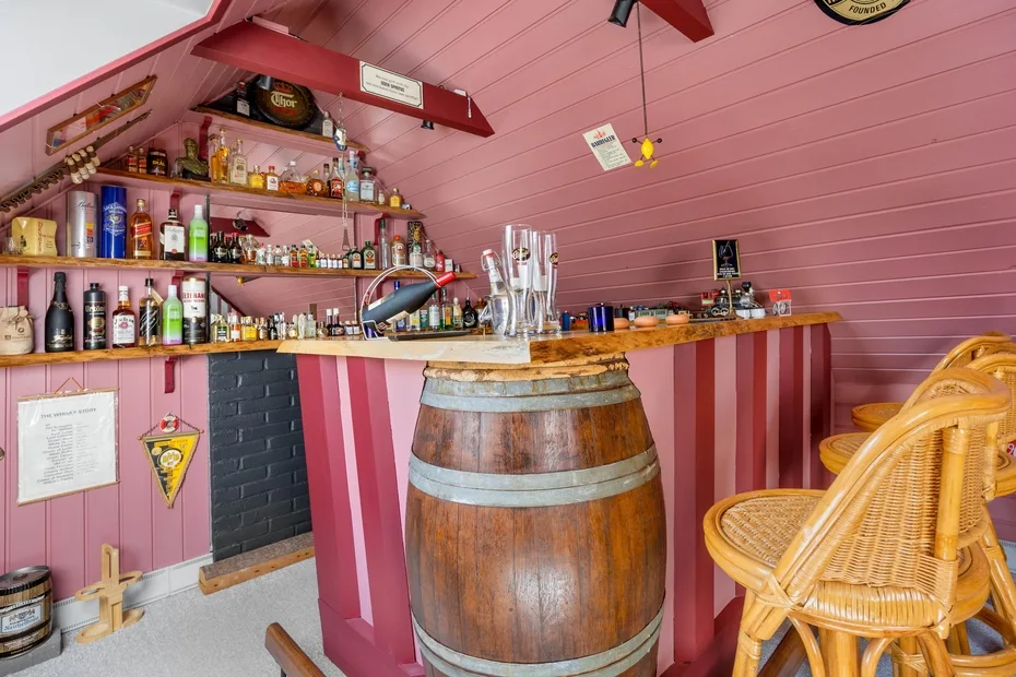 A compact bar with pink walls, stocked shelves, and a wooden barrel at the counter.