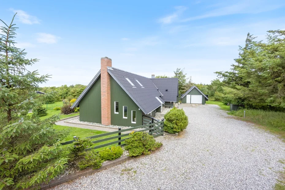A green house with a steep roof stands beside a gravel driveway and trees.