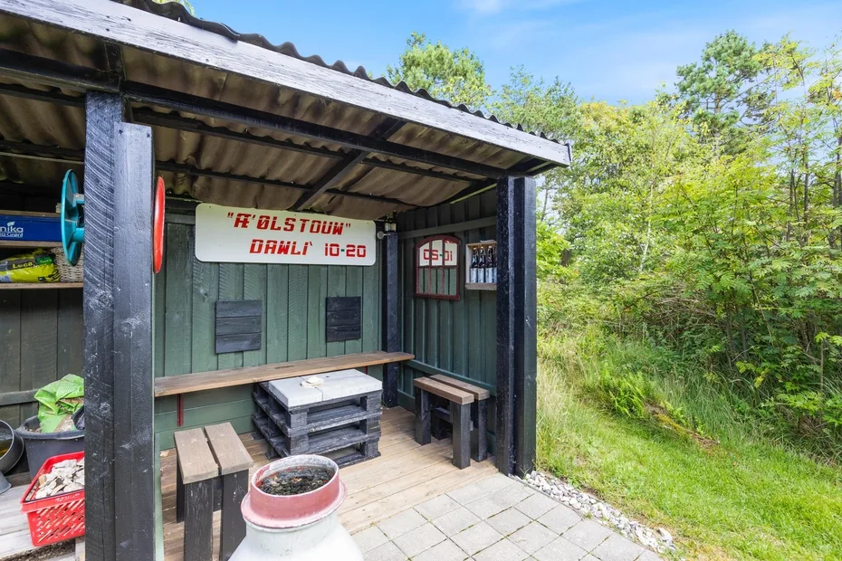A covered outdoor seating area with benches and a wooden table beside greenery.