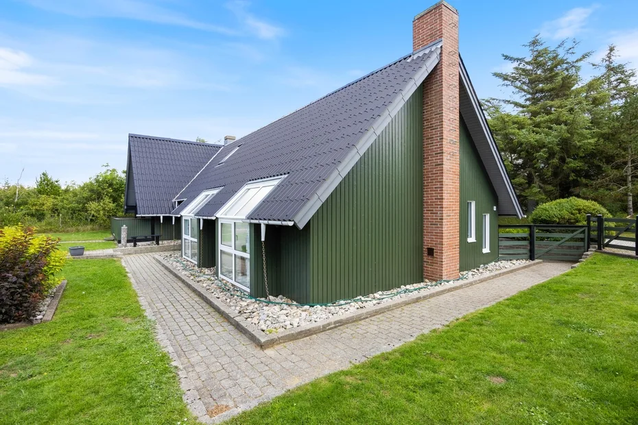 A green house with a steep roof and brick chimney stands beside a paved path.