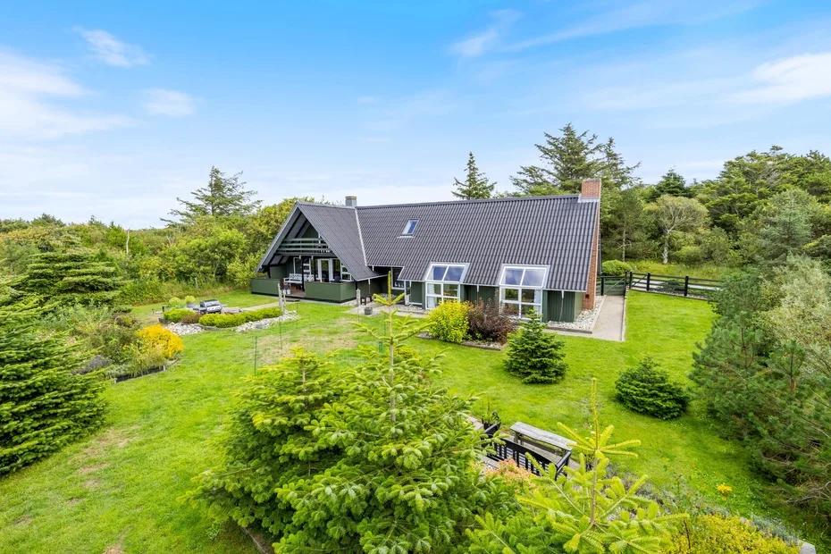 A house with a steep roof sits in a green garden surrounded by trees.