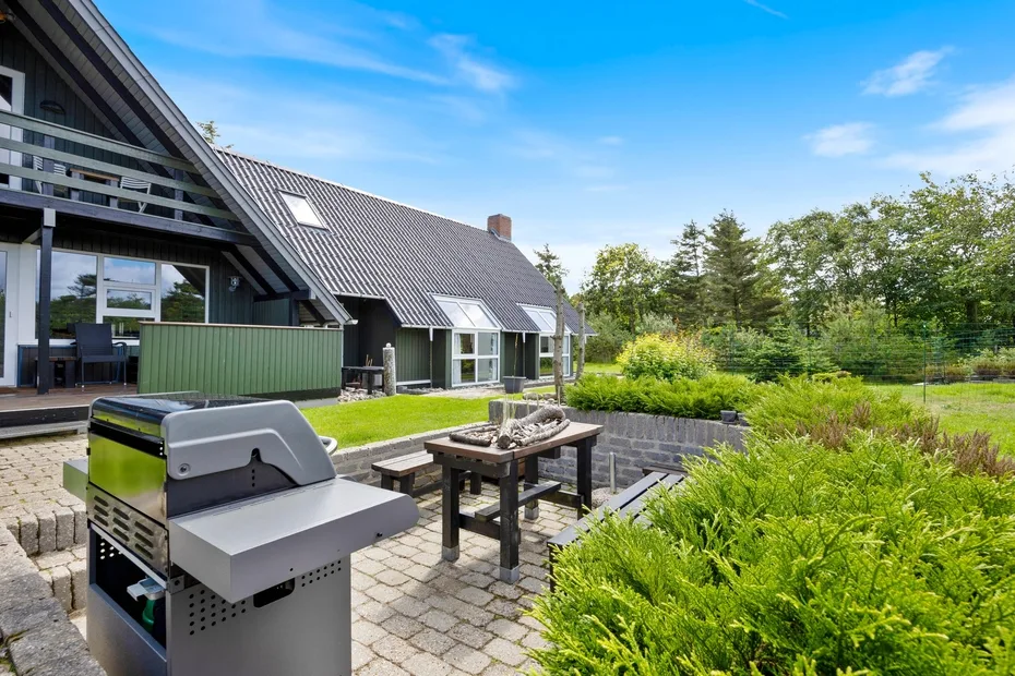A paved outdoor patio with a grill and table beside a house and garden.