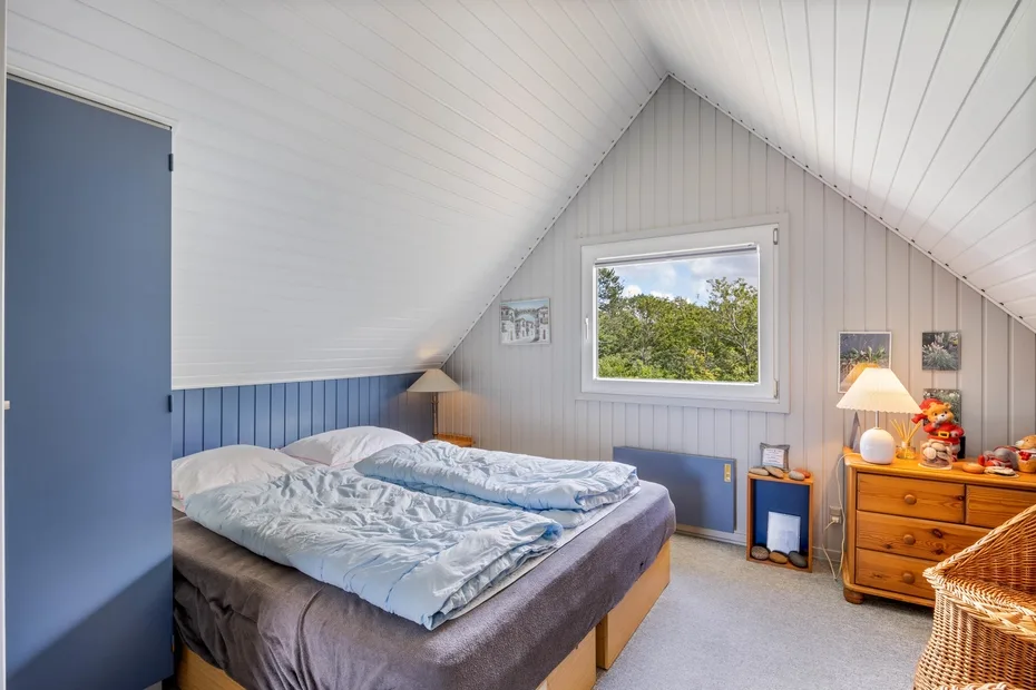 A bedroom with a bed beneath a sloped ceiling and a window overlooking trees.