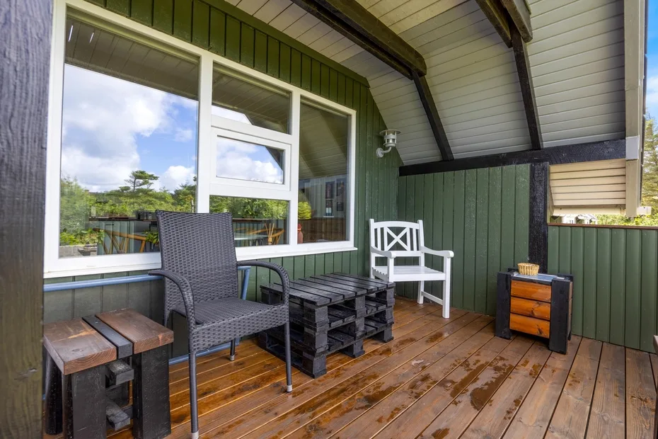 A covered wooden deck has outdoor seating beside large windows and green walls.