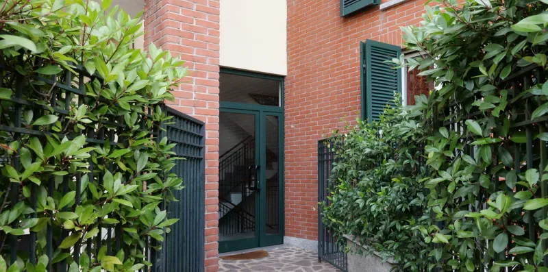 Entrada de un edificio de ladrillo enmarcada por setos frondosos y una reja metálica.