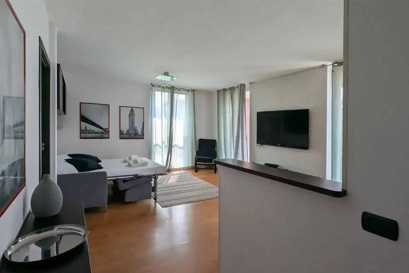 Una luminosa sala de estar tiene una cama, asientos y un televisor en la pared.