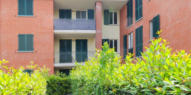 A brick apartment building with balconies and greenery in the foreground.