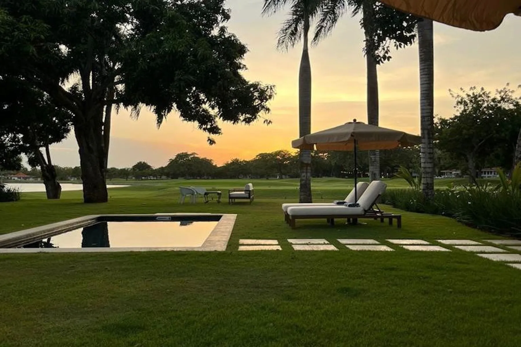 A landscaped garden beside a reflecting pool, with loungers beneath an umbrella at sunset.