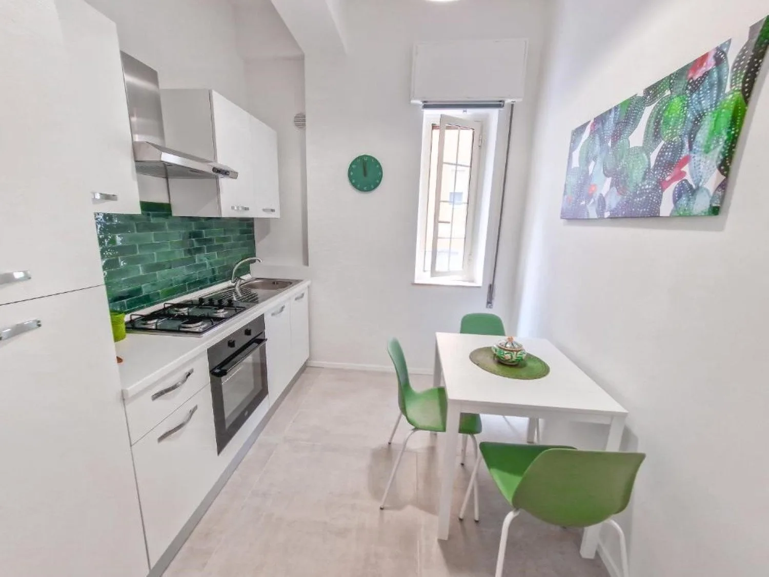 Una cocina estrecha con zona de comedor, gabinetes blancos, sillas verdes y una mesa pequeña.