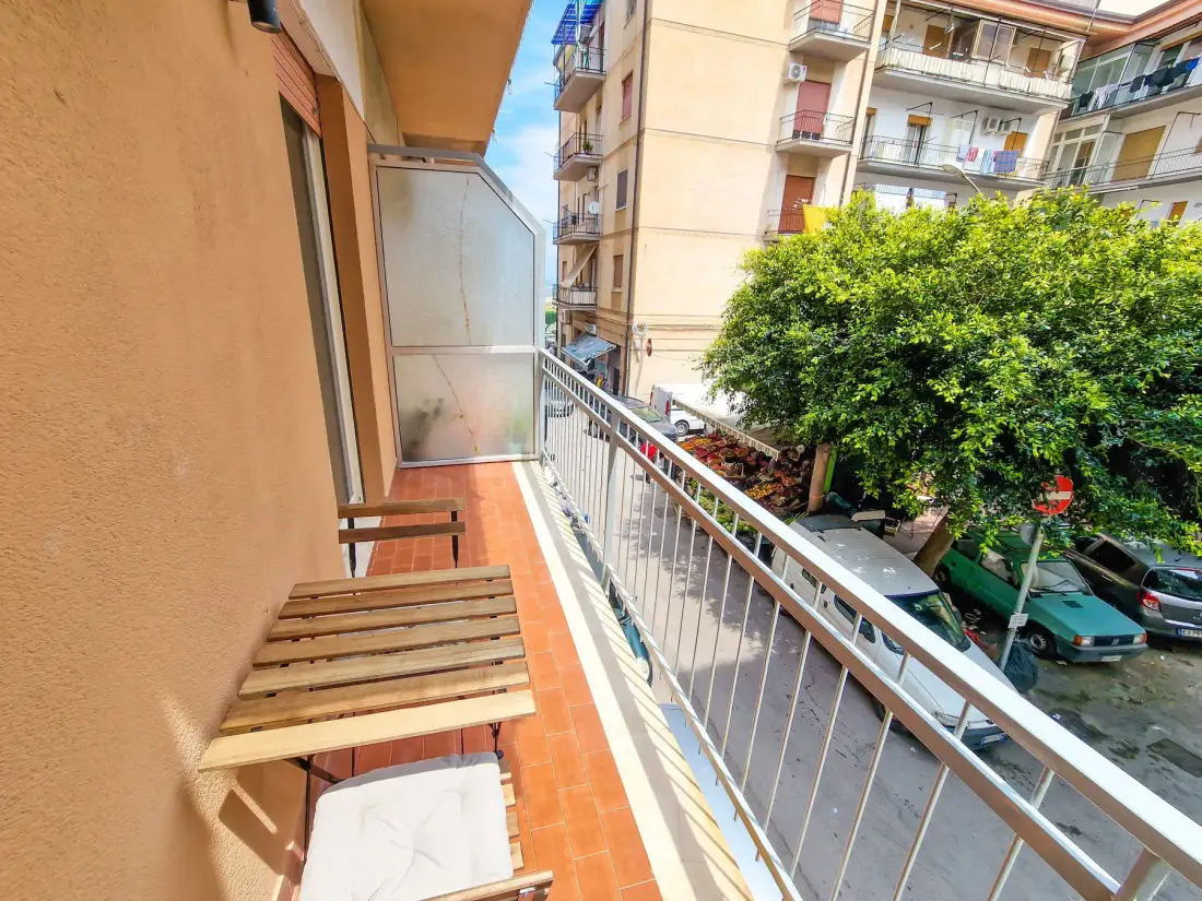 Un balcón estrecho con suelo de baldosas da a árboles frondosos y edificios de apartamentos cercanos.
