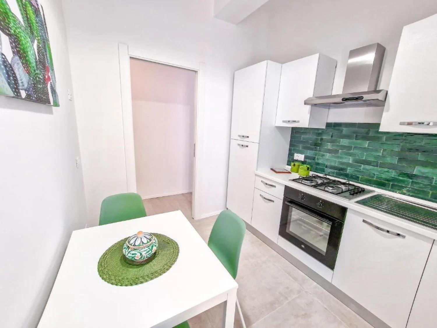 Una cocina luminosa con gabinetes blancos, un salpicadero verde y una pequeña mesa de comedor.