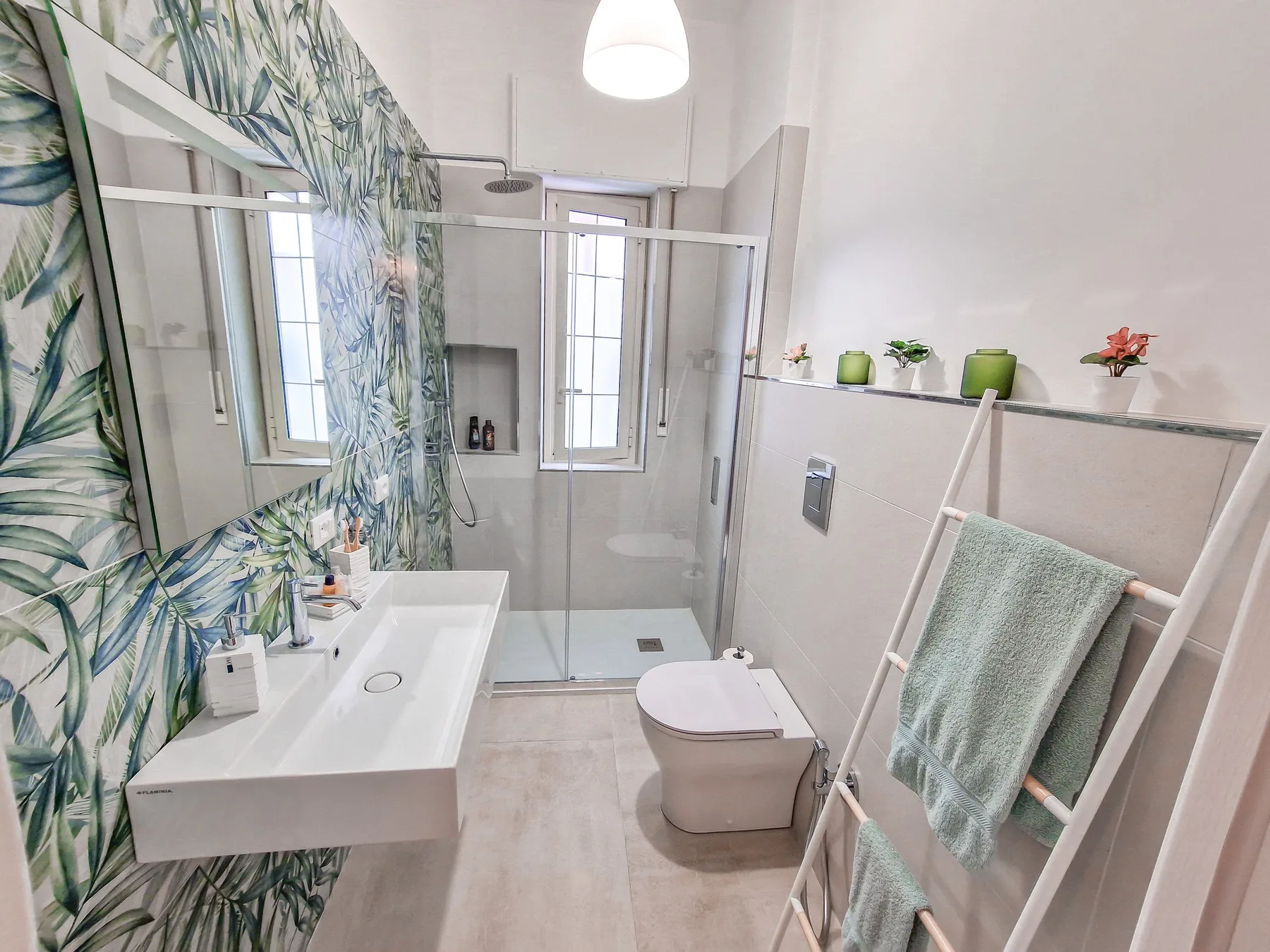 Un baño luminoso con ducha, lavabo blanco y revestimiento de pared con hojas.