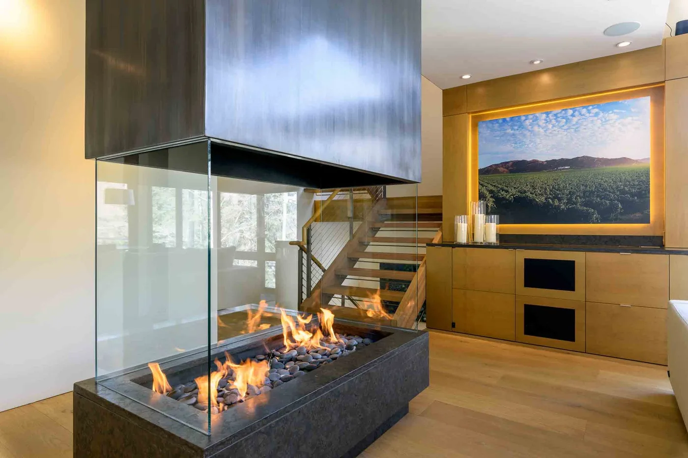 Una sala moderna muestra una chimenea acristalada junto a una escalera y una pantalla en la pared.