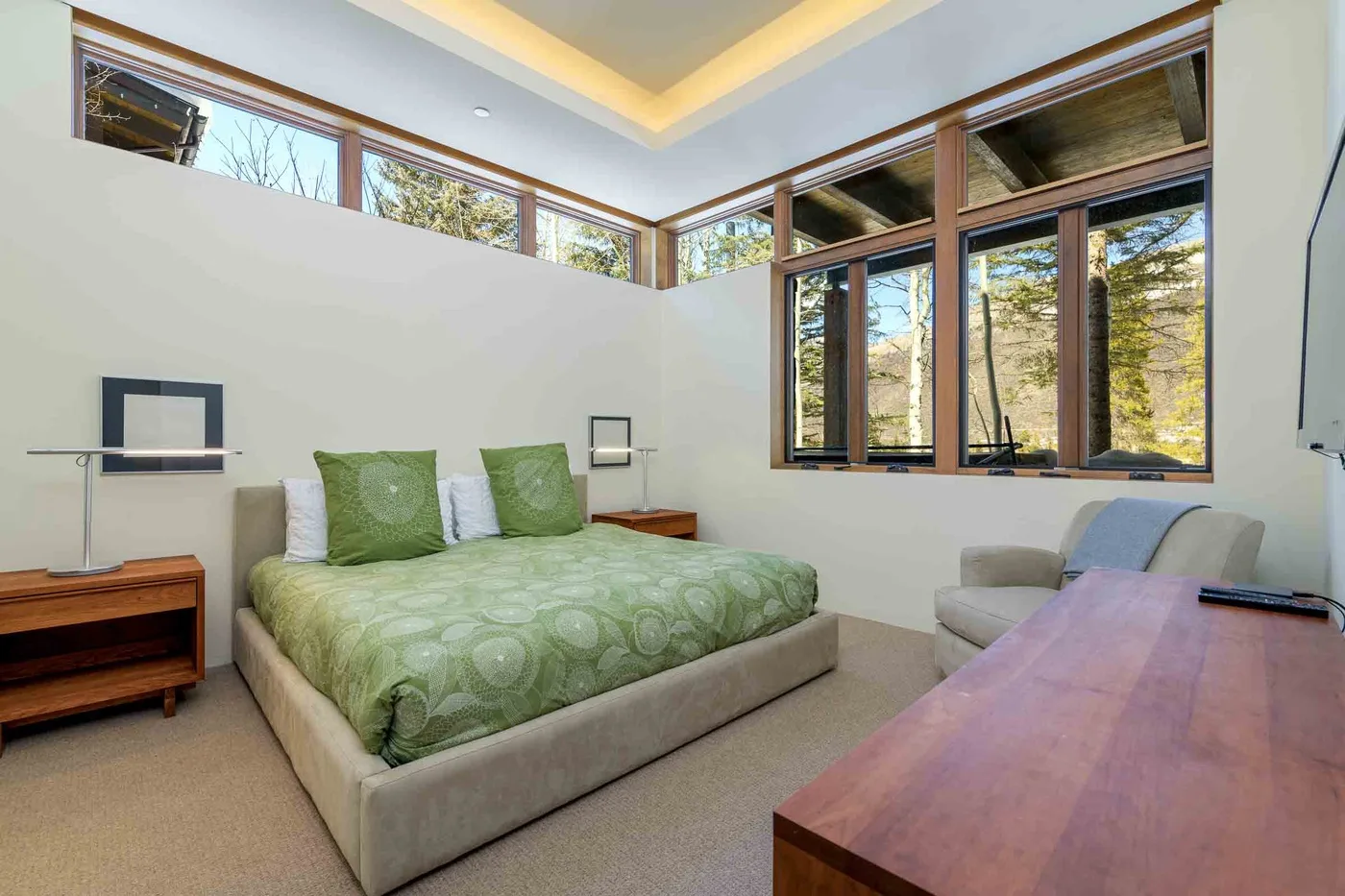 Un dormitorio con una cama verde, mesas de noche y grandes ventanas con vista a los árboles.