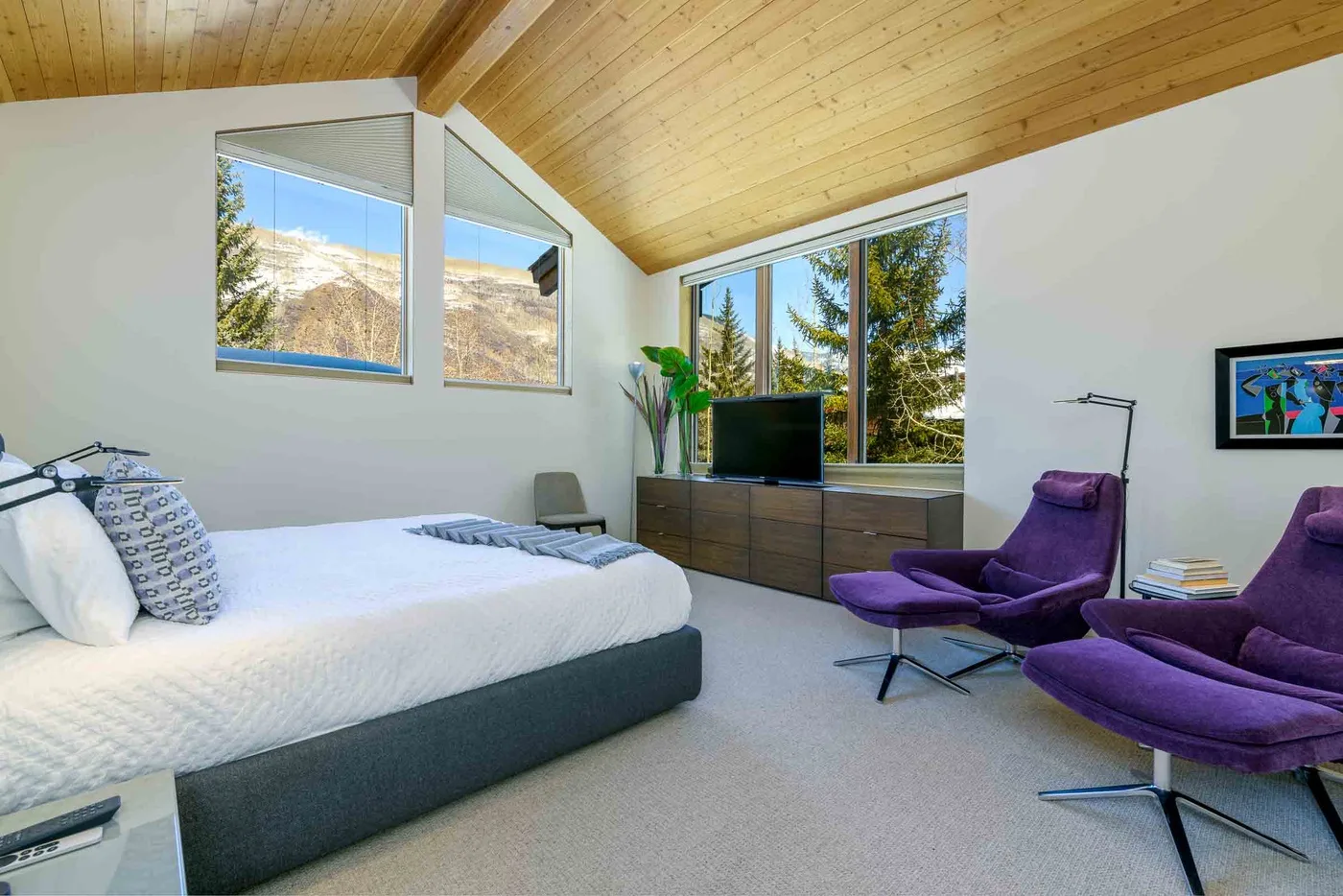 Un dormitorio con una cama grande, sillones morados y ventanas con vistas a los árboles.