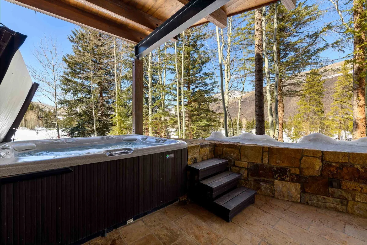 Una bañera de hidromasaje exterior cubierta está junto a un muro de piedra, con árboles al fondo.