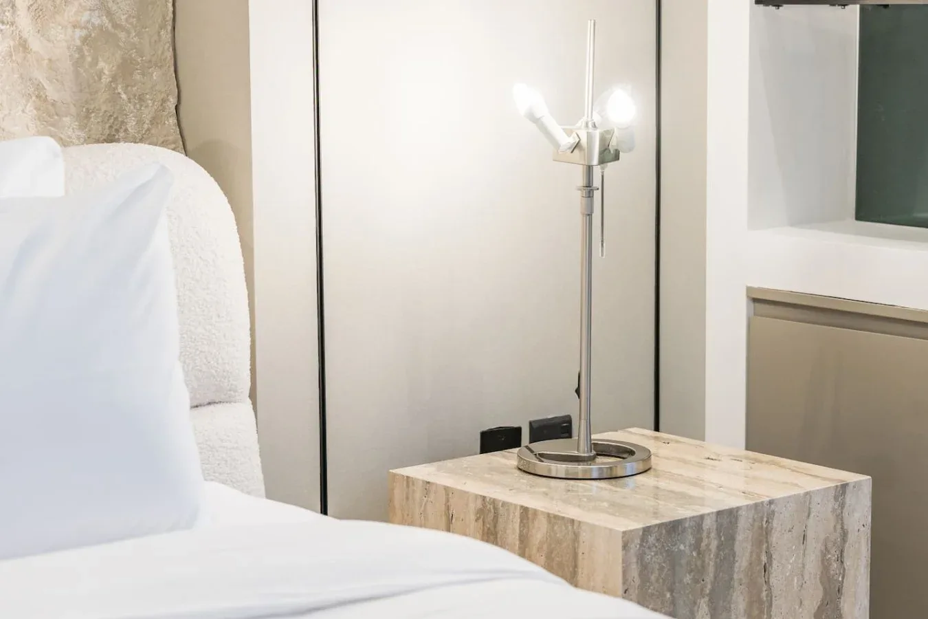 Dormitorio con ropa de cama blanca junto a una mesa auxiliar de textura pétrea y una lámpara.