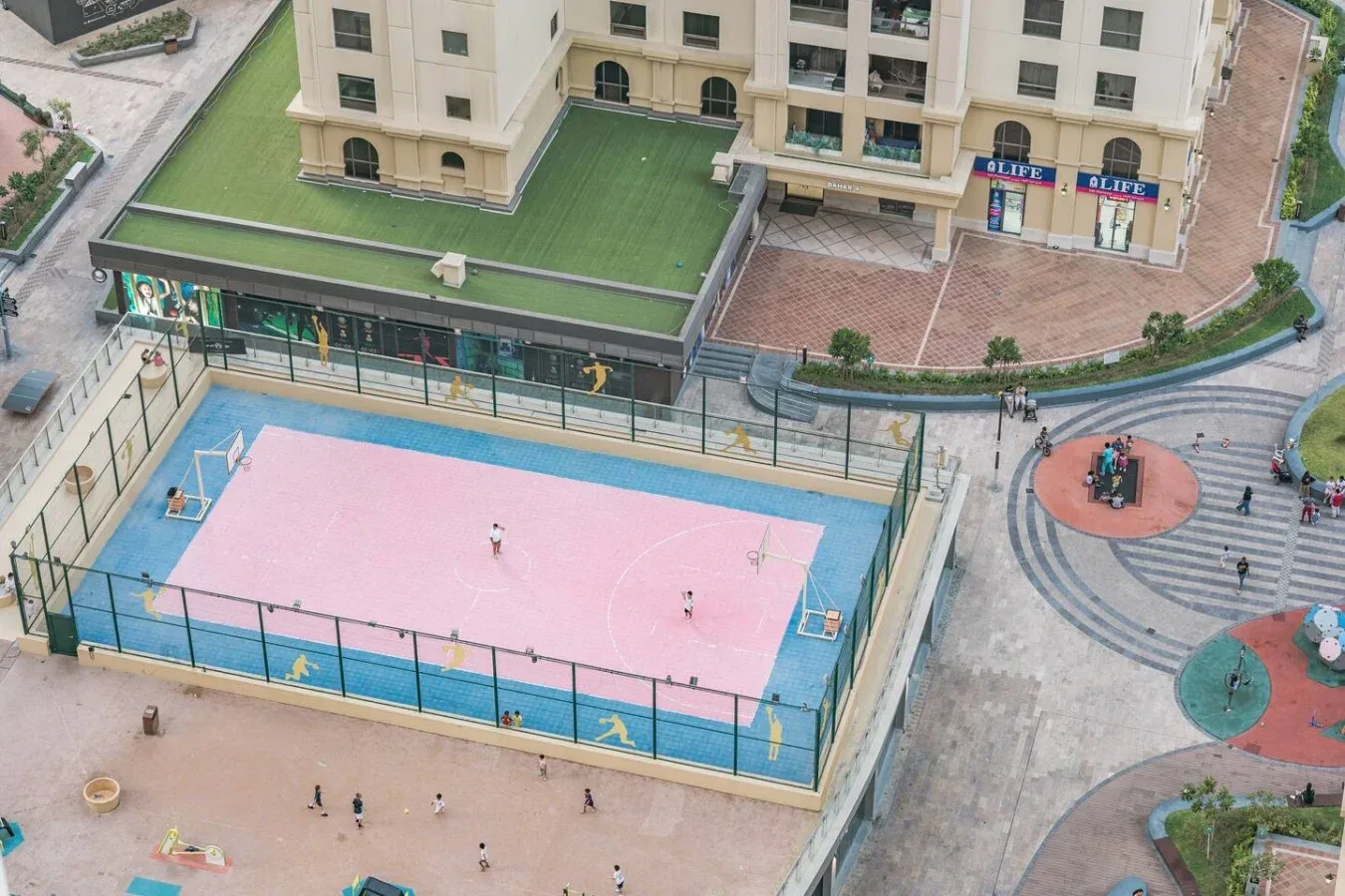 Una cancha de baloncesto al aire libre con superficie rosa está junto a una plaza pavimentada.
