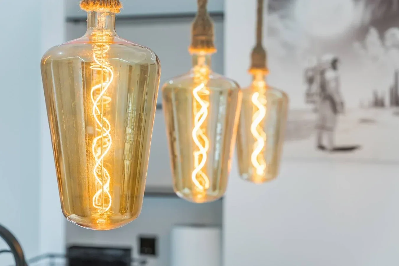 Tres lámparas colgantes iluminadas cuelgan frente a una pared clara y una obra enmarcada.