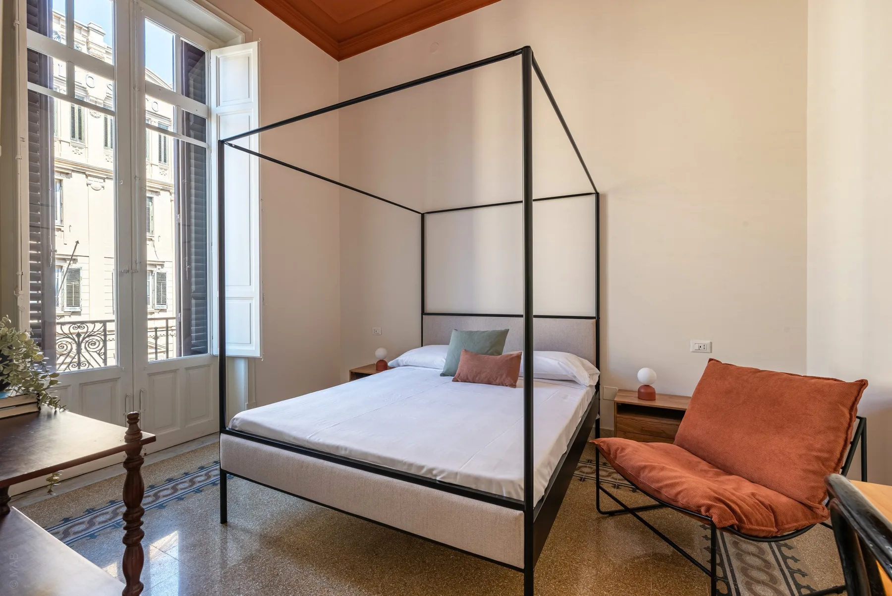 A bedroom with a canopy bed, tall windows, and an orange lounge chair.
