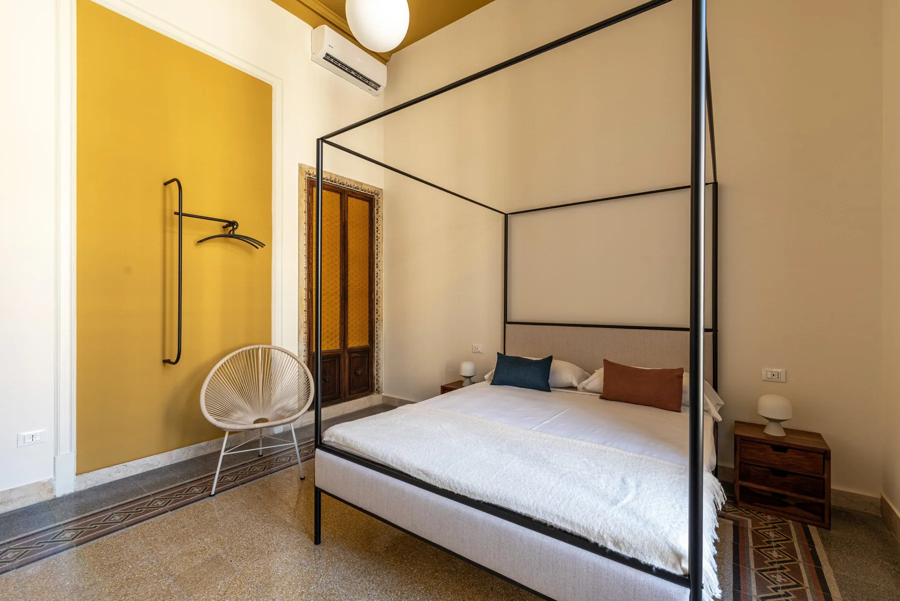 A bedroom with a bed beneath a tall black frame and a yellow accent wall.