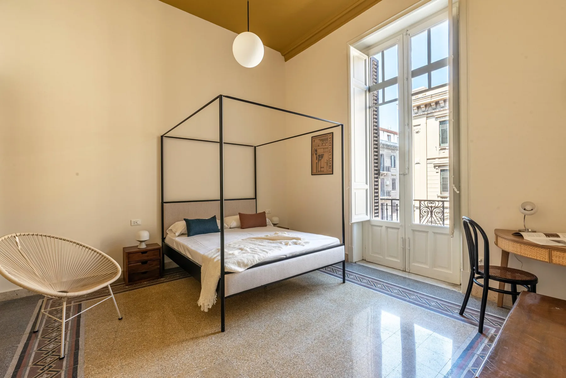 A bright bedroom has a large bed beneath a tall window.