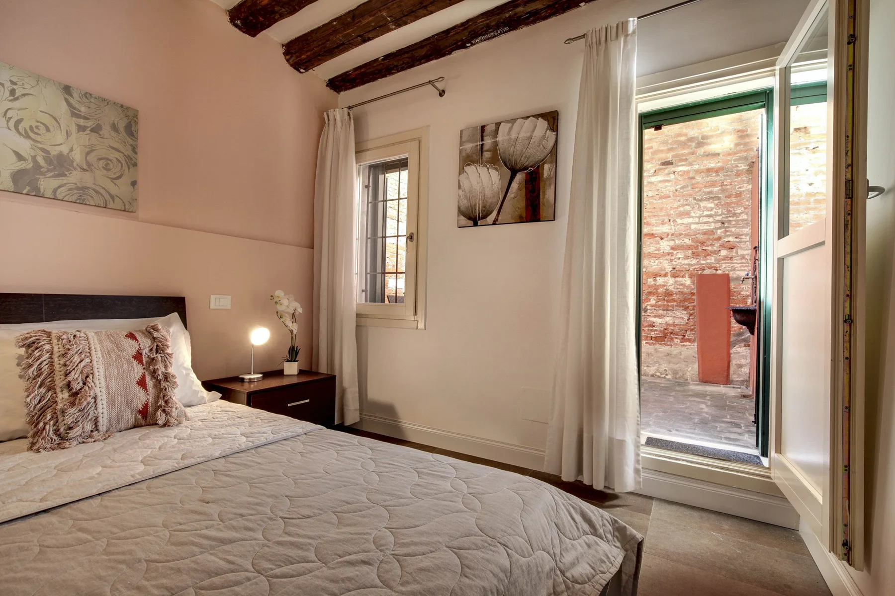 A bedroom with a bed, bedside table, and an open window overlooking a brick courtyard.