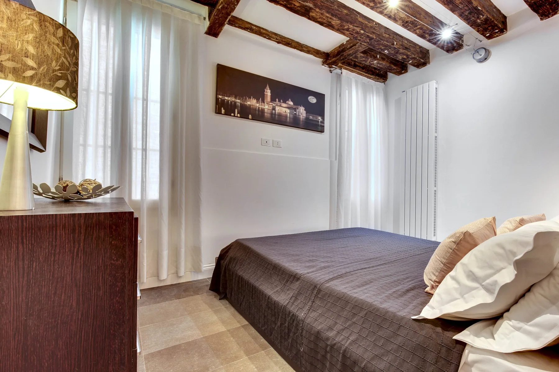 A bedroom with a double bed, curtained window, and exposed ceiling beams.