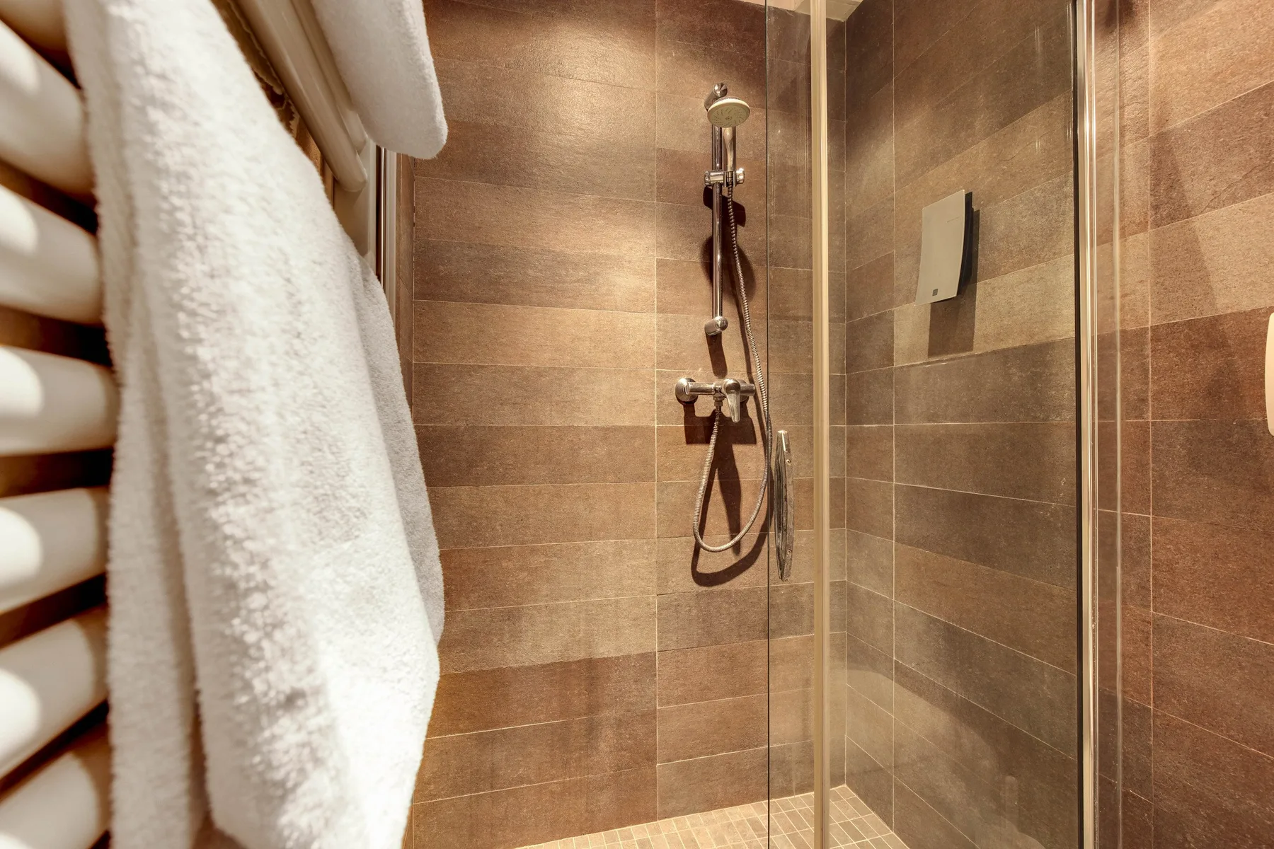 A tiled shower with a handheld showerhead and a white towel hanging nearby.