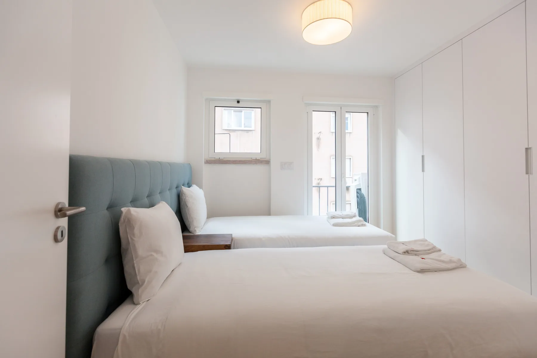 Un dormitorio luminoso con dos camas, ropa de cama blanca y un cabecero azul tapizado.