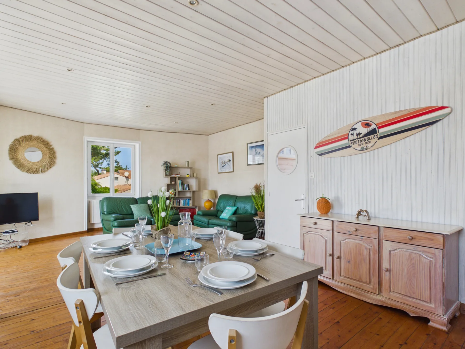 Dining area with a set table beside green seating and a wall-mounted surfboard.