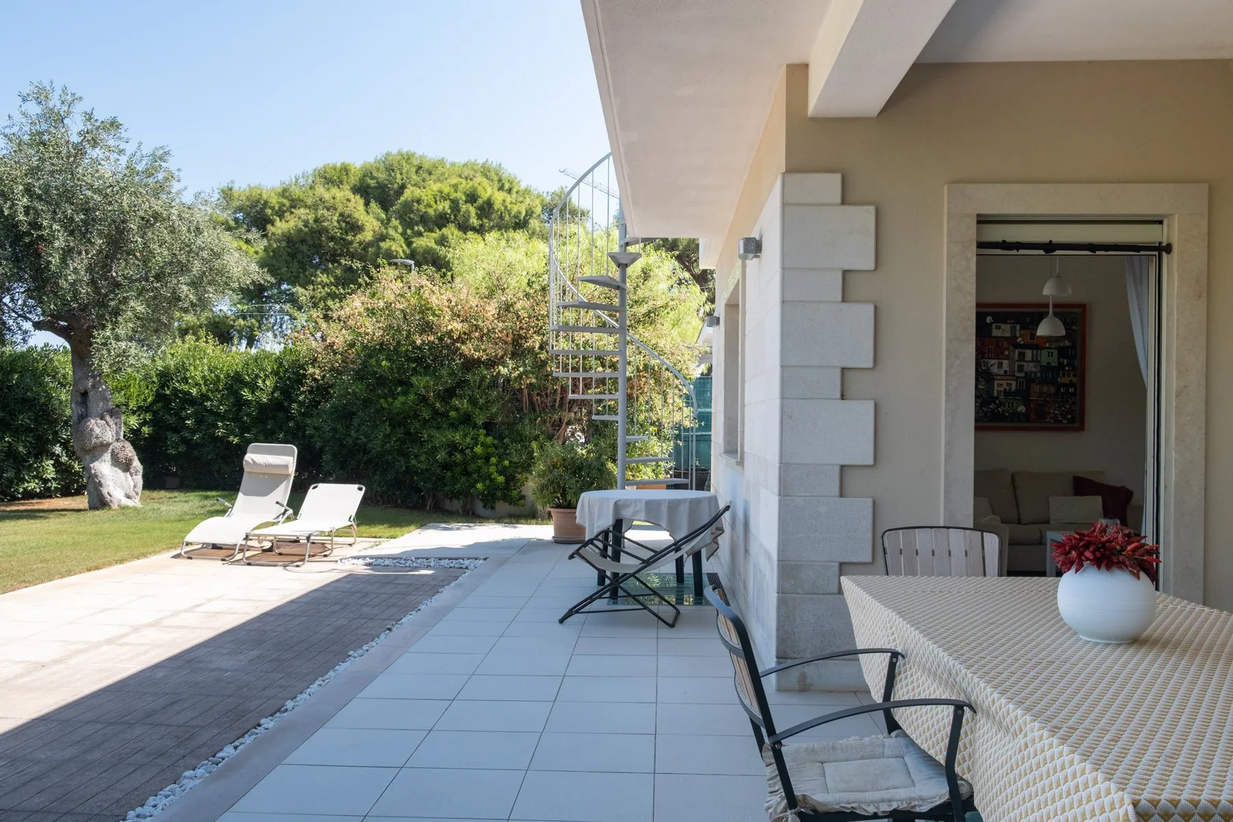 A sunny patio beside a house has outdoor seating and a leafy garden.