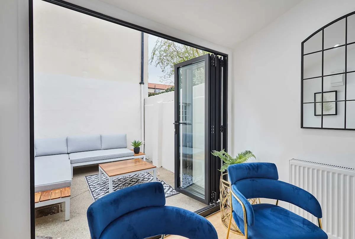 Una sala luminosa con un sofá gris, asientos azules y puertas de vidrio abiertas.