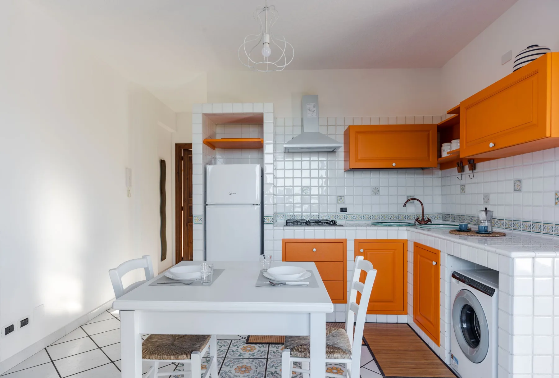 Una cocina luminosa con armarios naranjas, una mesa blanca y paredes de azulejos.