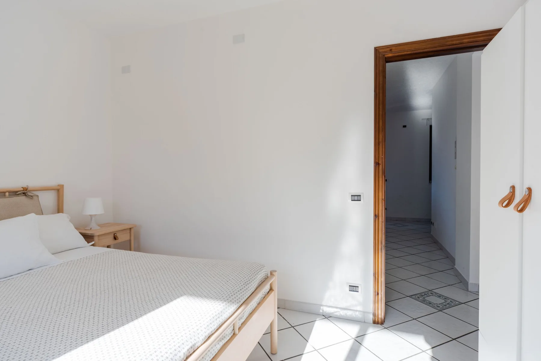 Un dormitorio luminoso con una cama y una puerta abierta a un pasillo de baldosas.