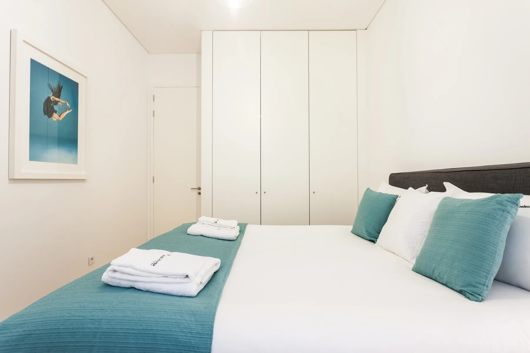 A bedroom with a white bed, teal pillows, and a folded towel.