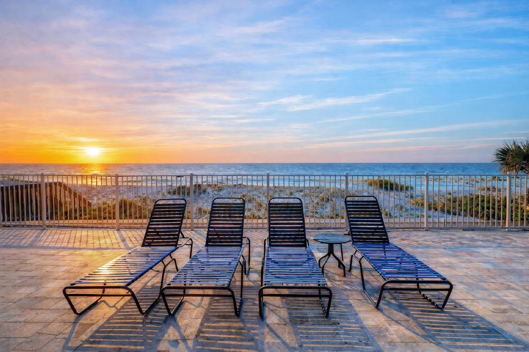 A seaside terrace with reclining seating overlooks the ocean at sunset.