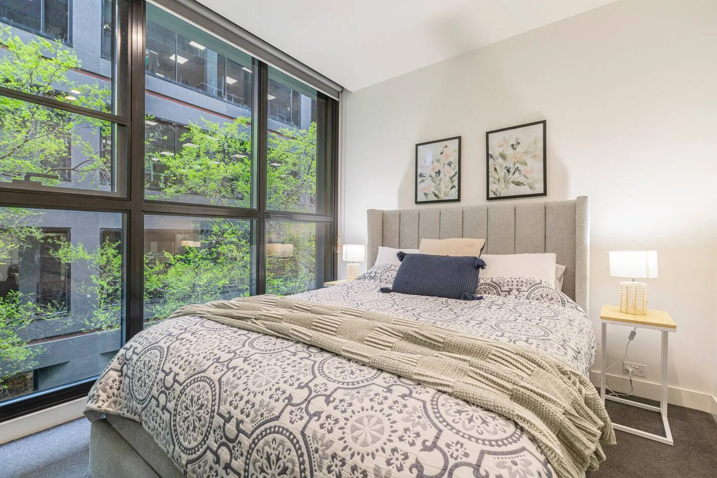 A bedroom with a large bed, patterned bedding, and leafy views through wide windows.