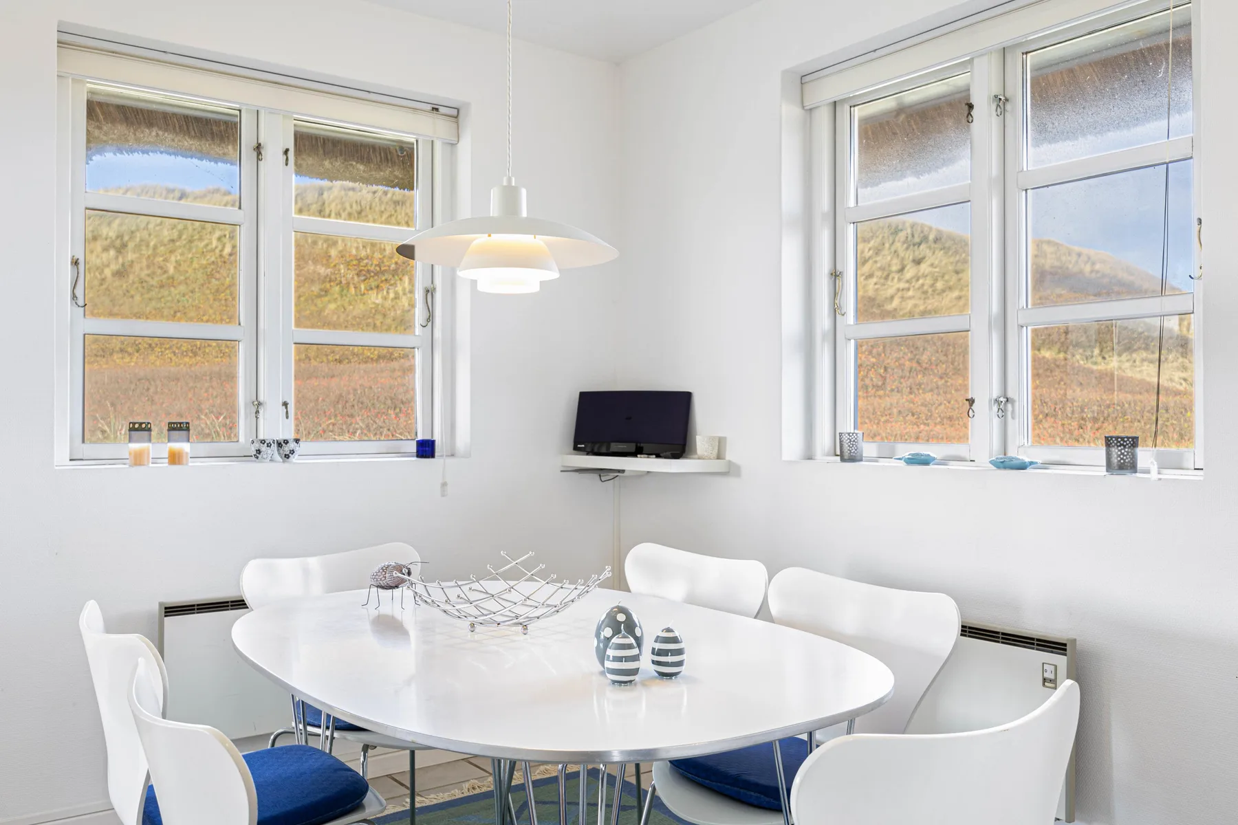 Un comedor luminoso tiene una mesa blanca y grandes ventanas con vistas al paisaje.