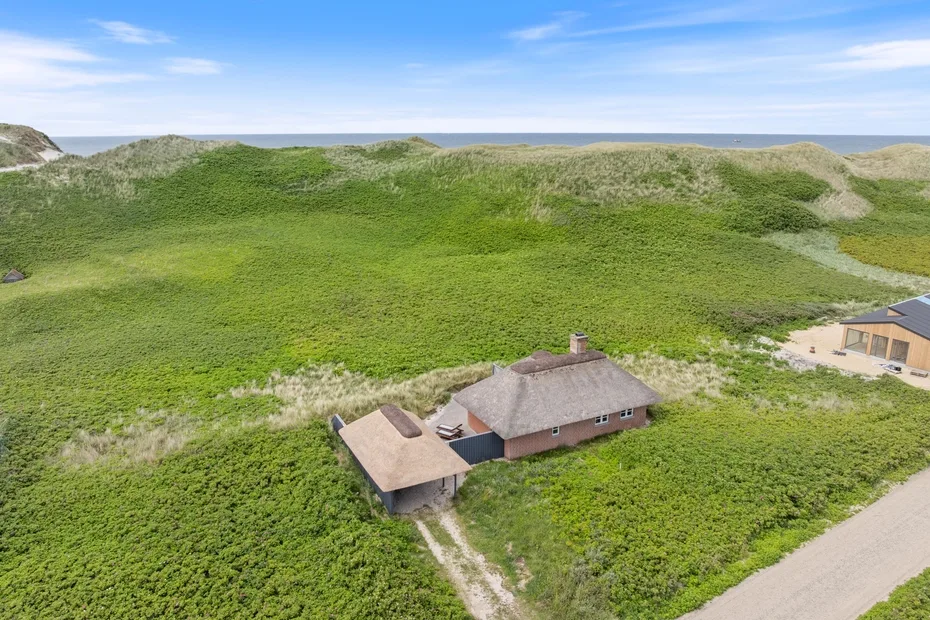 Vista aérea de una casa entre dunas verdes, con el mar en el horizonte.