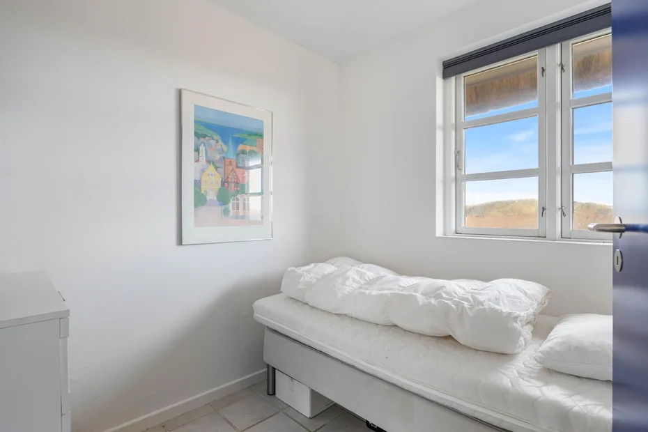 Un dormitorio luminoso con una cama blanca bajo una ventana.